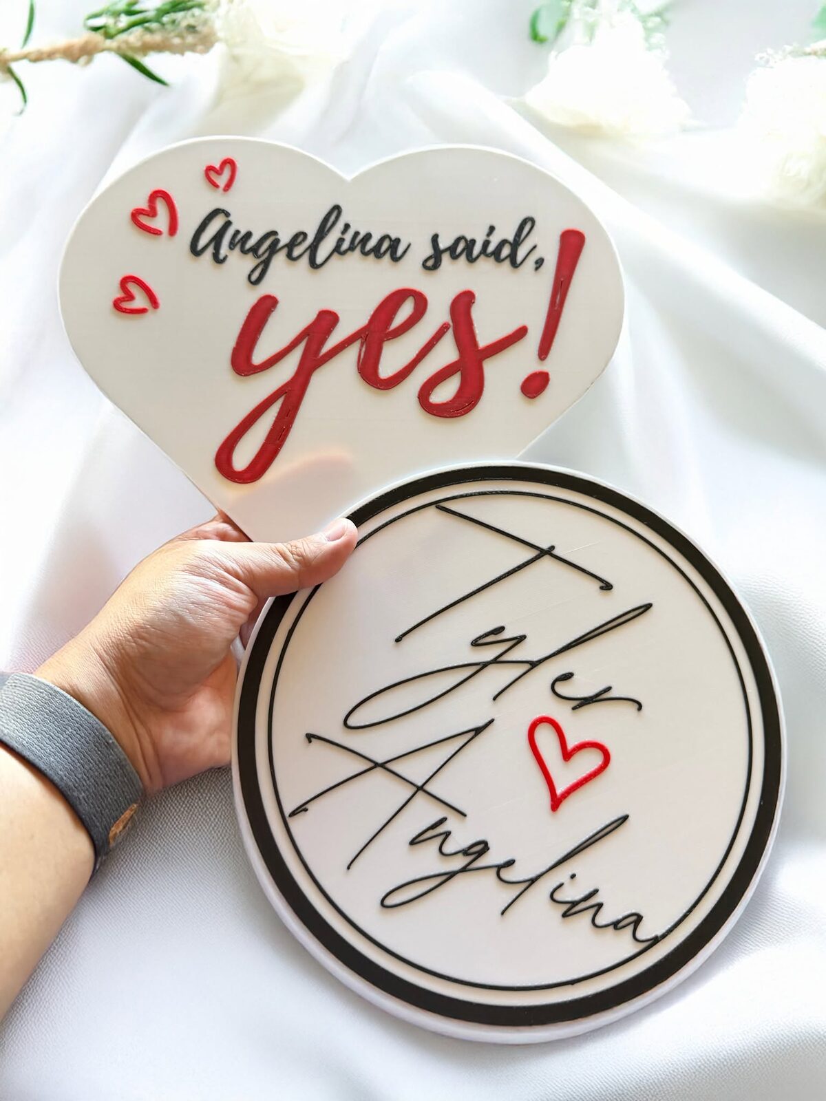 A hand holding two finished custom props — a heart reading "Angelina said, yes!" and a round sign with the couple's names